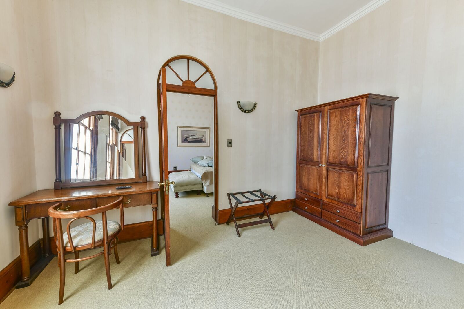 A room with a wooden desk, chair, and wardrobe, a mirror, and a bed in the background.