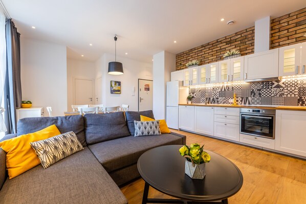 A modern living room with a grey sofa, yellow cushions, a black coffee table with a vase of flowers, and a kitchen with white cabinets and patterned backsplash.