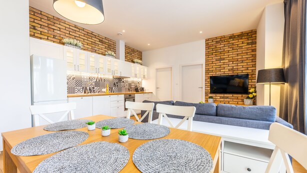 Ein modernes Wohnzimmer mit grauem Sofa, einem an einer Ziegelwand montierten Fernseher und einem Essbereich mit Holztisch und grauen Untersetzern.