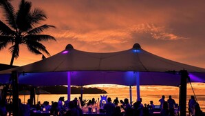 Evento en la playa con un gran toldo, palmeras y una puesta de sol al fondo.