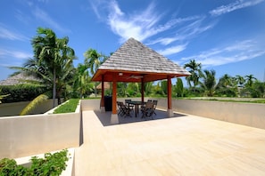 A covered outdoor seating area with a table and chairs, surrounded by a low wall and palm trees.