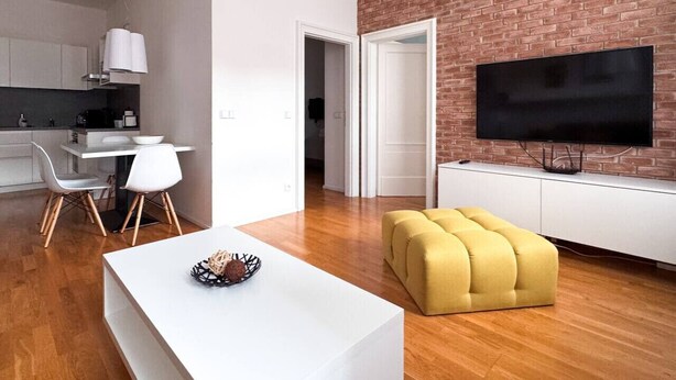 Un salon moderne avec une table à manger blanche, un pouf jaune et un téléviseur fixé sur un mur en briques.