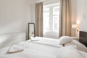 A hotel room with two beds, a bedside table with a lamp, a large window with curtains, and a mirror on the wall. - Riverside area apartment near the Dancing house! (Prague 2)