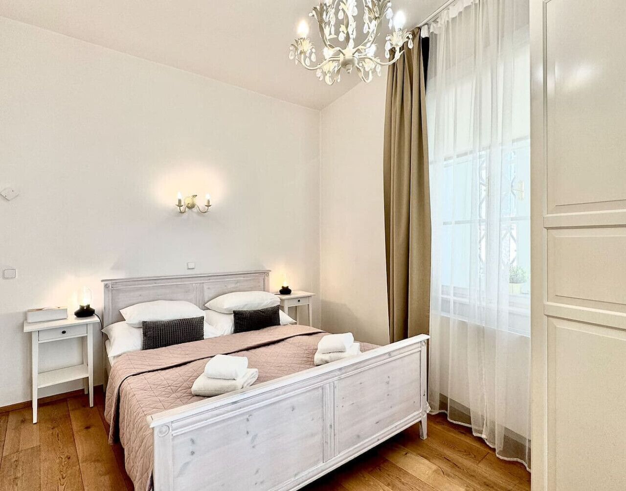 A bedroom with a bed, nightstands, a chandelier, and a window with curtains.