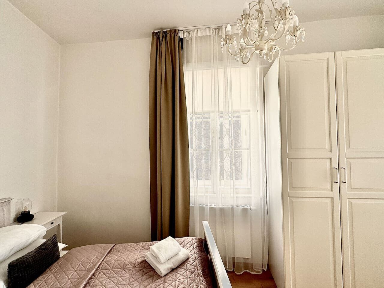 Een slaapkamer met een bed, nachtkastje, dressoir, een raam met dunne gordijnen en een kroonluchter.