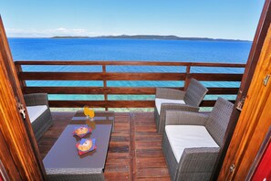 Eine Holzterrasse mit Korbmöbeln und einem Tisch, mit Blick auf das klare, blaue Meer und sanfte Hügel in der Ferne.