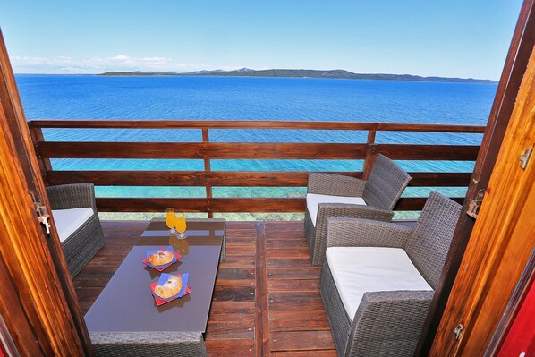 Una terraza de madera con muebles de mimbre y una mesa, con vistas a un mar azul y colinas distantes.