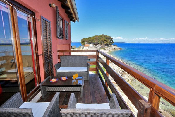 A balcony with wicker furniture, a table with a glass, and a view of the sea and a small island.