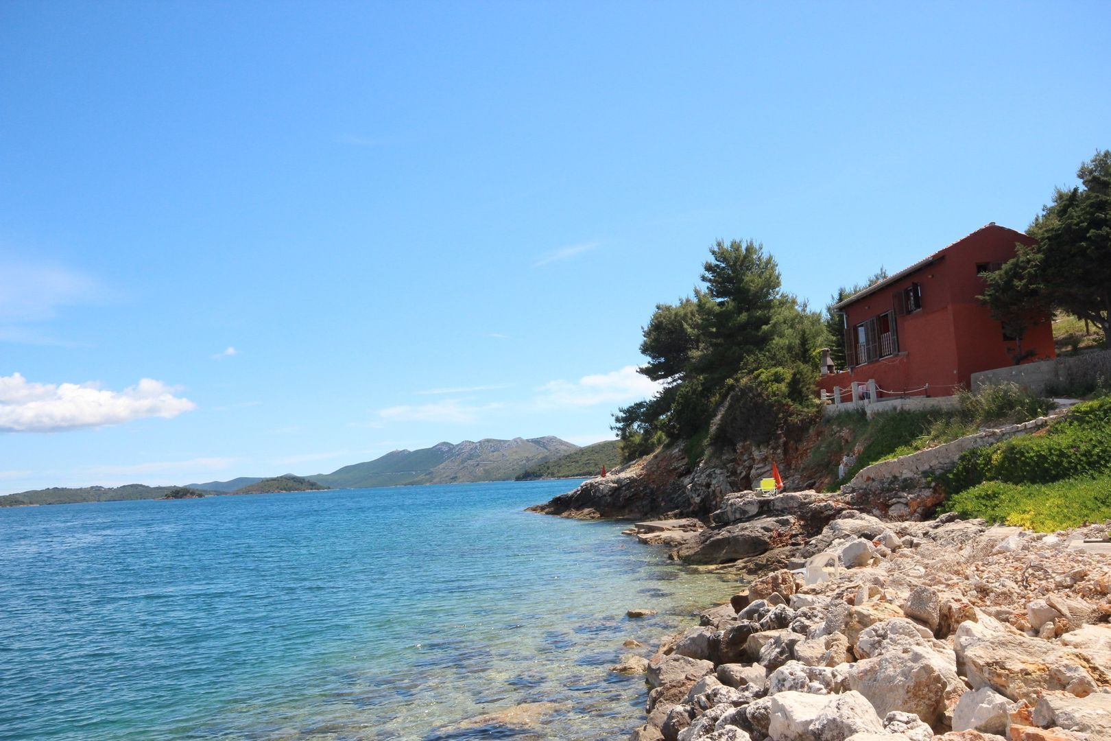 A coastal property with a clear view of the sea, rocky shoreline, and a red building with a balcony.