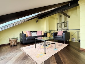 Un salon de style industriel, avec un plafond haut, des poutres en bois foncé, un canapé en cuir noir, une table basse en verre et un mur jaune.
