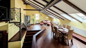 A spacious loft with a high ceiling, exposed wooden beams, a dining area with a long table and chairs, a staircase with a stained glass window, and a cozy seating area with cushions.