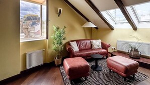 A loft-style room with a red leather sofa, a matching ottoman, a floor lamp, and a large window overlooking a cityscape.