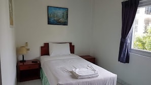 A single bed with white linens, a nightstand with a lamp, a painting on the wall, and a window with curtains. - Welcome Inn Hotel Karon Beach (Karon)