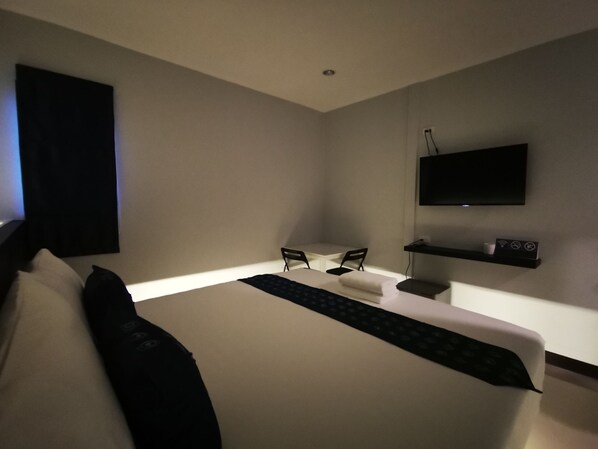 Habitación de hotel moderna con una cama grande, un televisor de pantalla plana en la pared, una pequeña mesa con dos sillas y un estante con cafetera.