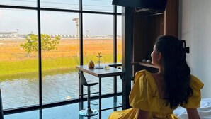 A room with large windows overlooking a field, a small table with a chair, and a television mounted on the wall. - Skyline Resort (Bang Phli)