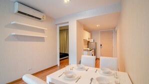 Un espace repas moderne avec une table blanche dressée pour quatre personnes, un climatiseur fixé au plafond et une vue sur la chambre adjacente.