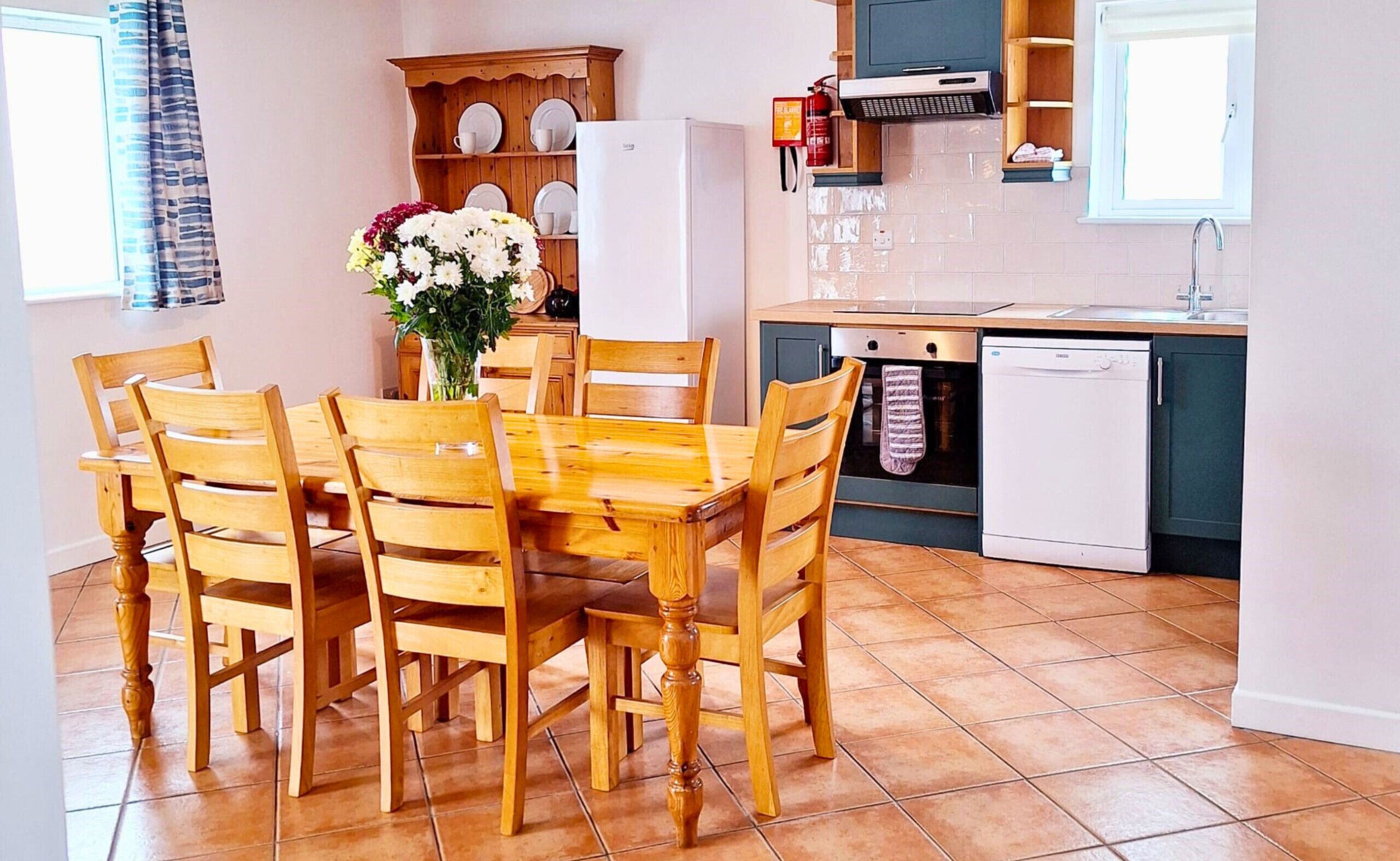 A compact kitchen with a dining table, chairs, a refrigerator, a dishwasher, and a wooden cabinet with dishes.