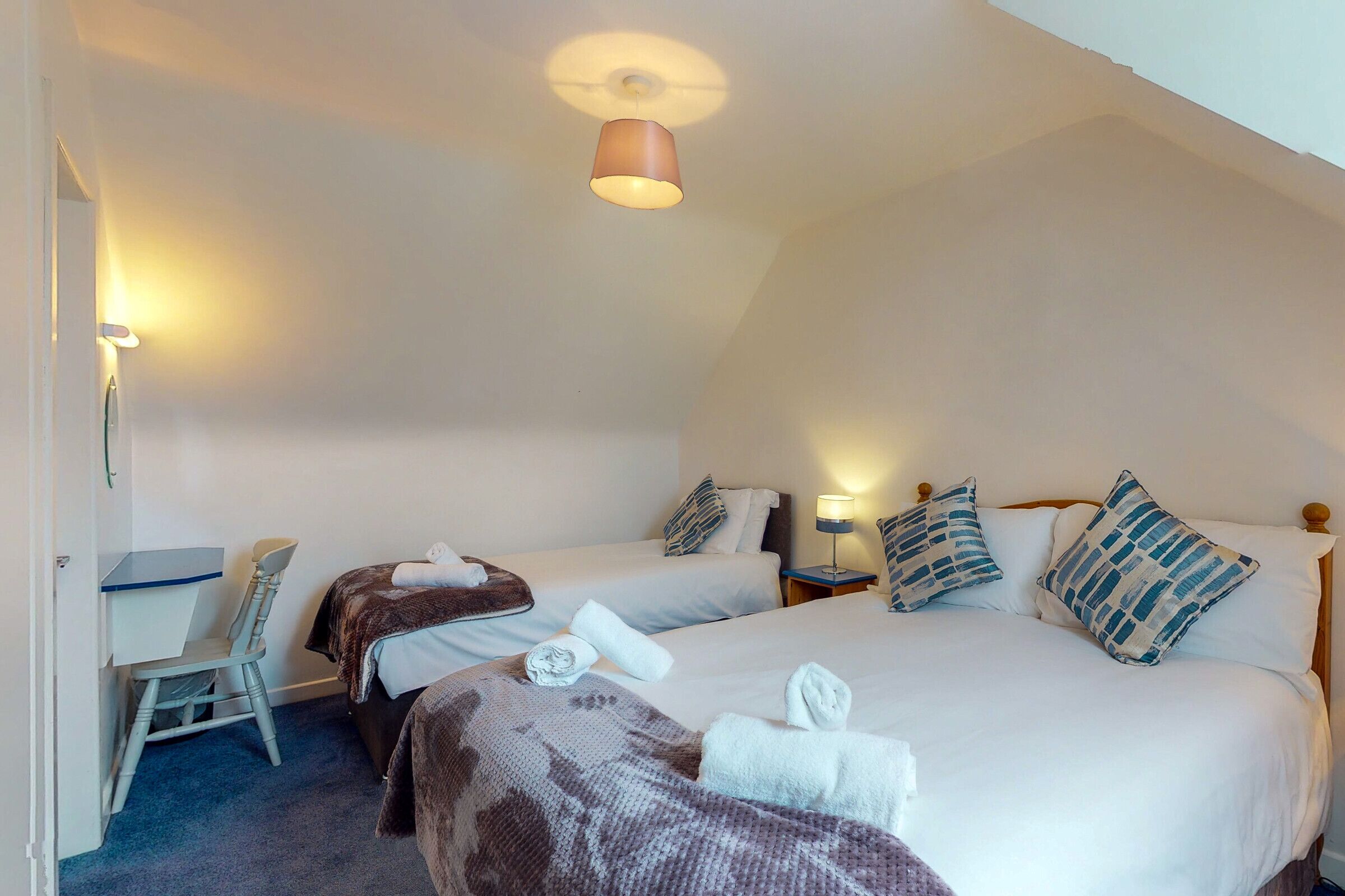 A hotel room with two beds, a desk, and a chair. There are towels on the beds and a lamp on the desk.
