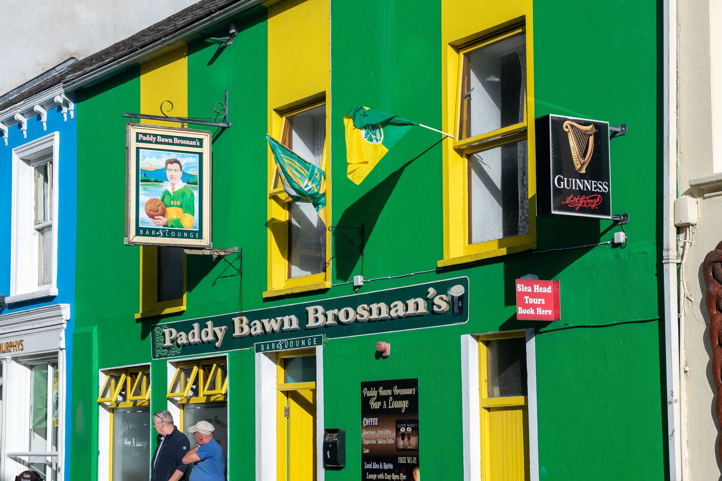 Een gebouw met een geelgroene gevel, met borden voor Paddy Bawn Brosnan's Bar & Lounge en Guinness.