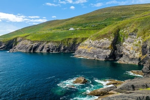 A coastal landscape with rocky cliffs, a clear blue sea, and a grassy hill with buildings. - Dingle Harbour Cottages (Dingle)
