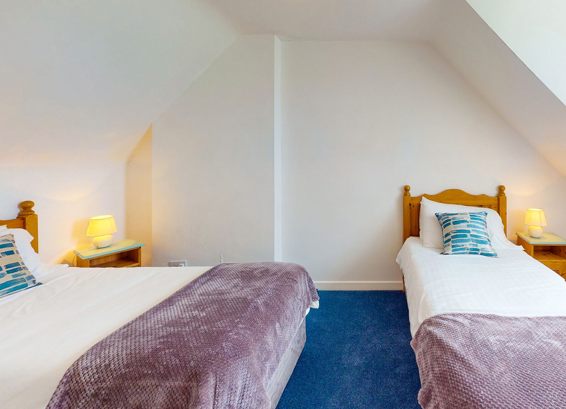 A room with two beds, each with a wooden headboard, a blue carpet, and bedside lamps.