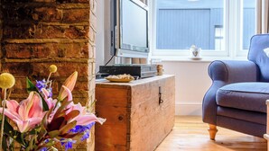 Un salón acogedor con chimenea de ladrillo, un televisor sobre un cofre de madera, un sillón azul y una ventana con vista a una cerca azul.