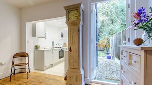 Una habitación amplia con piso de madera, un reloj de pie grande, una silla y vistas a una cocina y un jardín a través de una puerta abierta.
