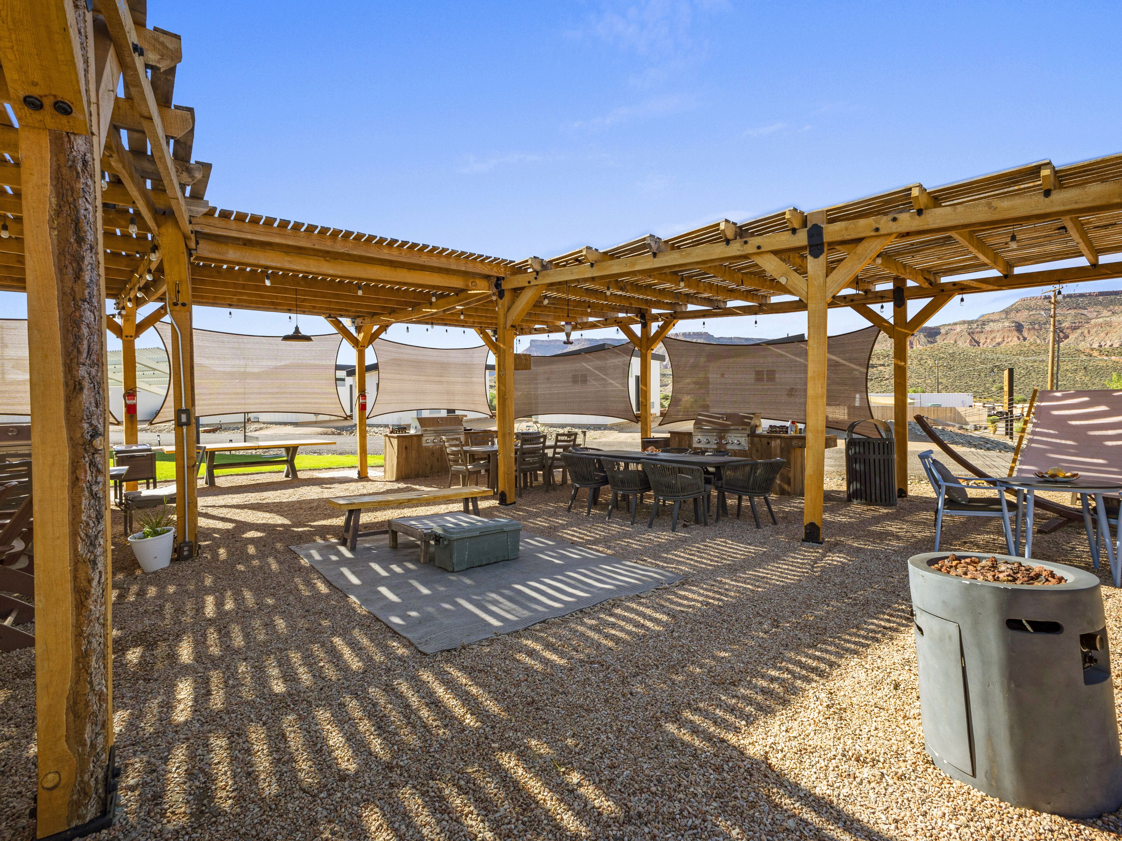 An outdoor patio area with wooden pergolas, tables, chairs, and a barbecue grill.