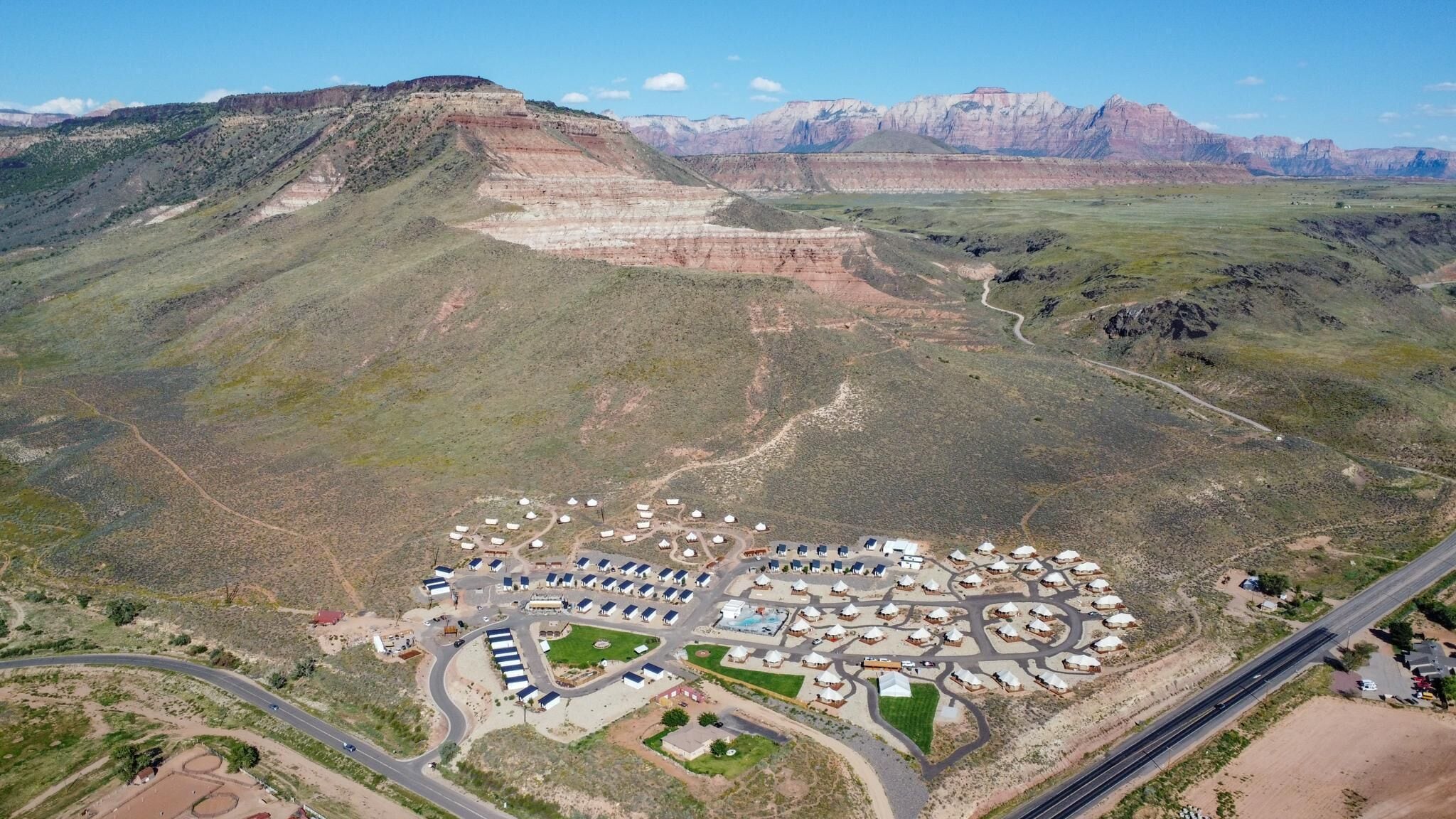 An aerial view of a resort with yurts arranged in a semi-circular pattern, surrounded by a paved area with a swimming pool and solar panels.