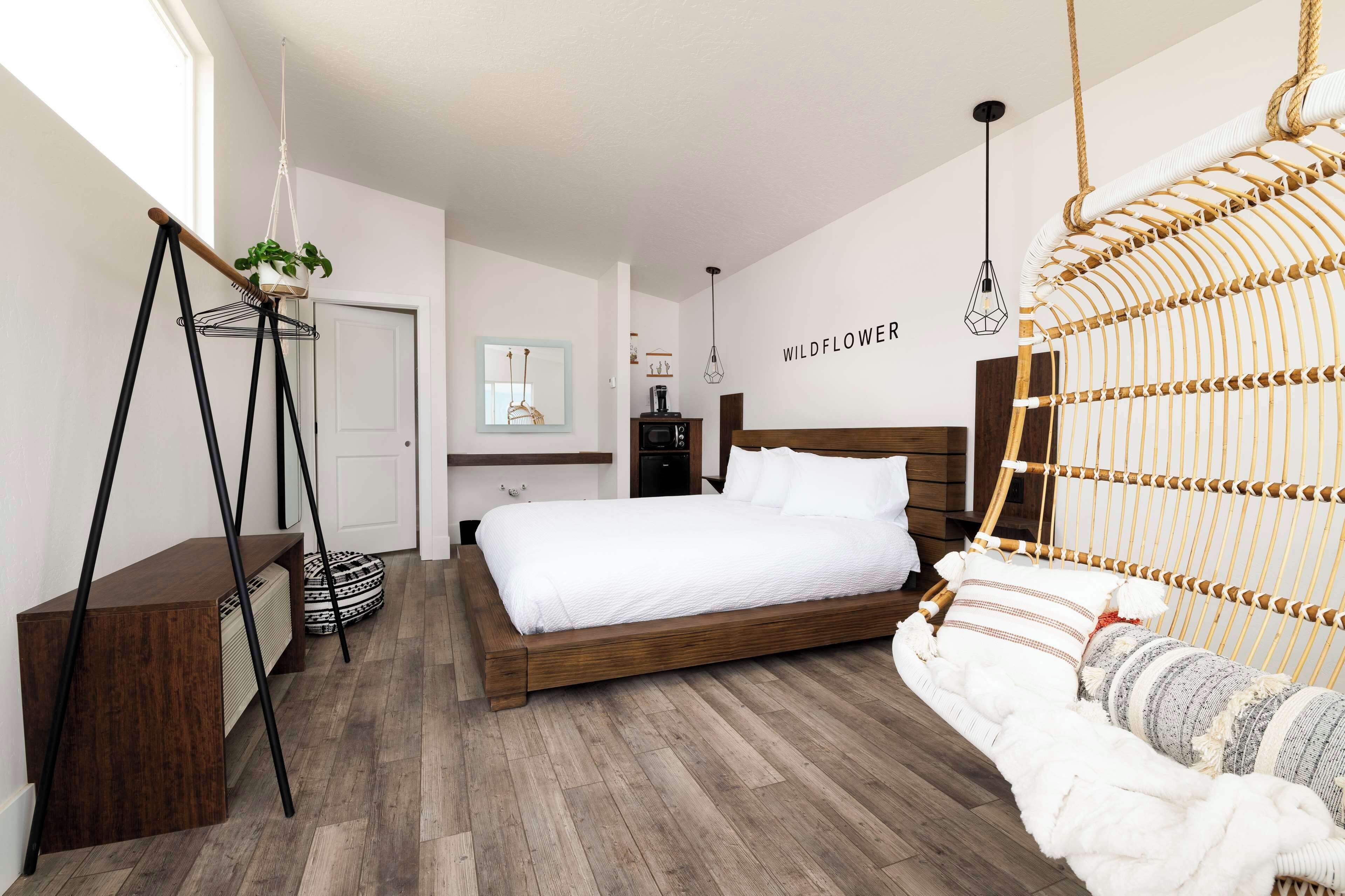 Ein modernes Schlafzimmer mit einem Holzbett, einem Hängekorbstuhl, einer kleinen Pflanze auf einem Sockel und einer wandmontierten Leuchte.