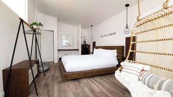 Ein modernes Schlafzimmer mit einem Holzbett, einem Hängekorbstuhl, einer kleinen Pflanze auf einem Sockel und einer wandmontierten Leuchte.