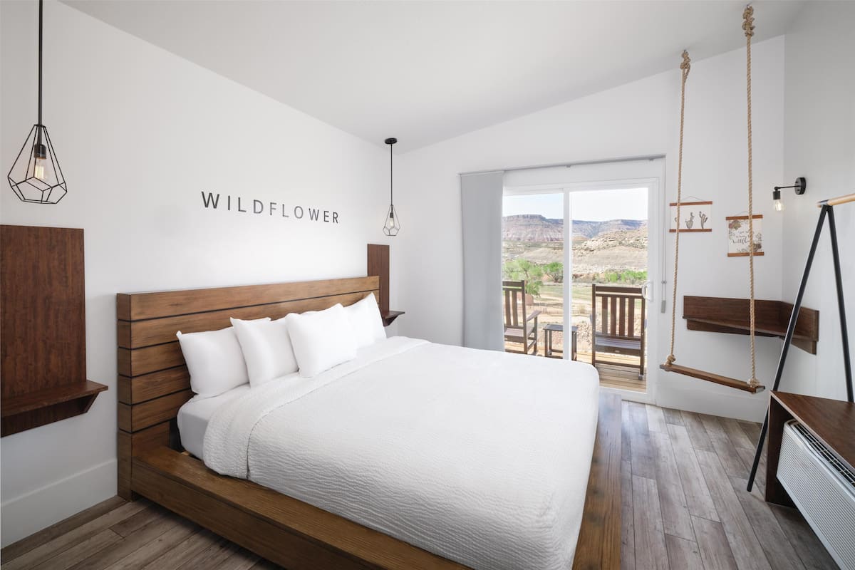 Quarto com cama de madeira, roupa de cama branca, vista para o exterior e luminária fixada na parede.