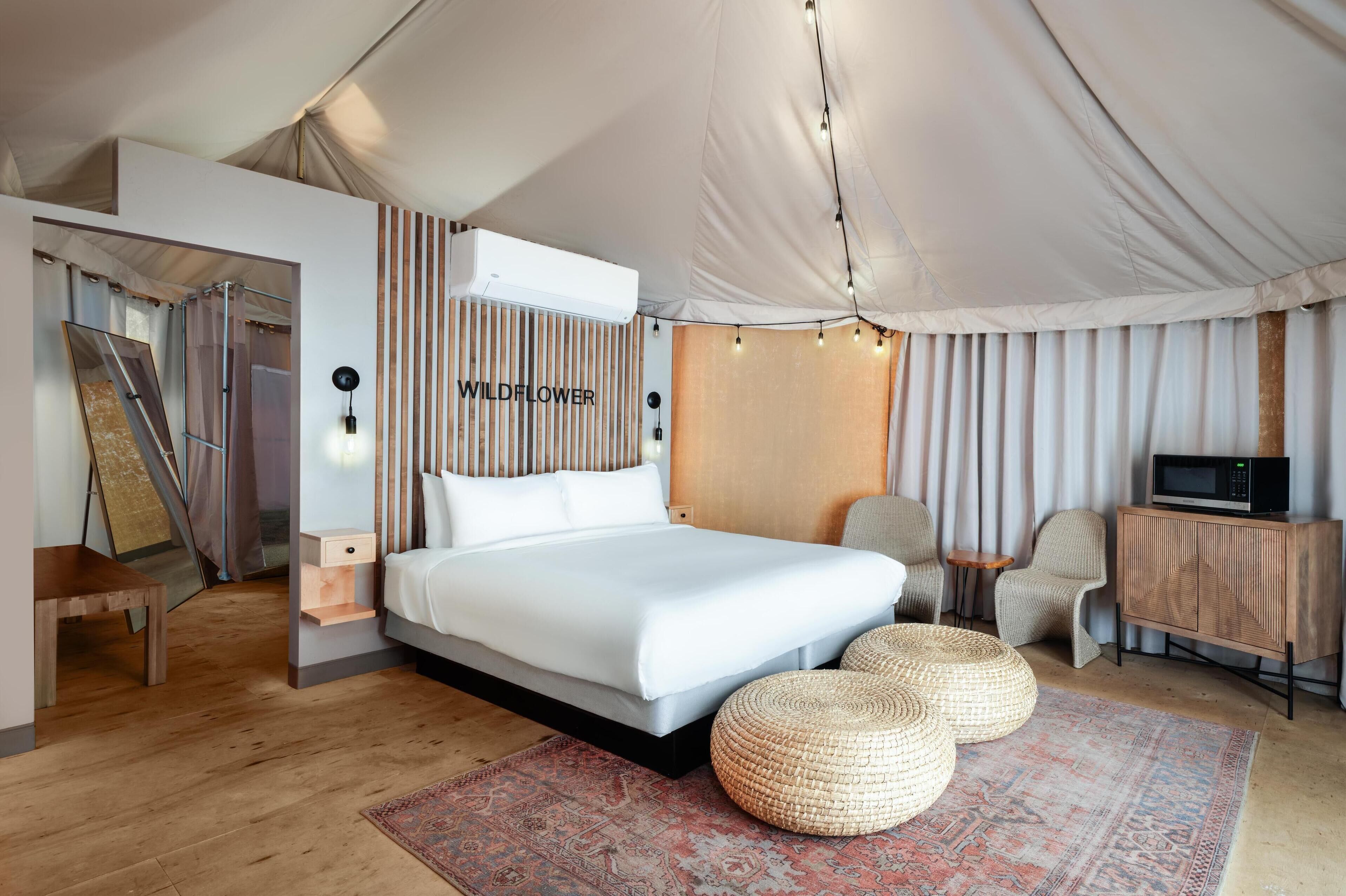 Une chambre à coucher moderne avec un grand lit, une table de chevet en bois, une petite table, un téléviseur et un pouf en osier.
