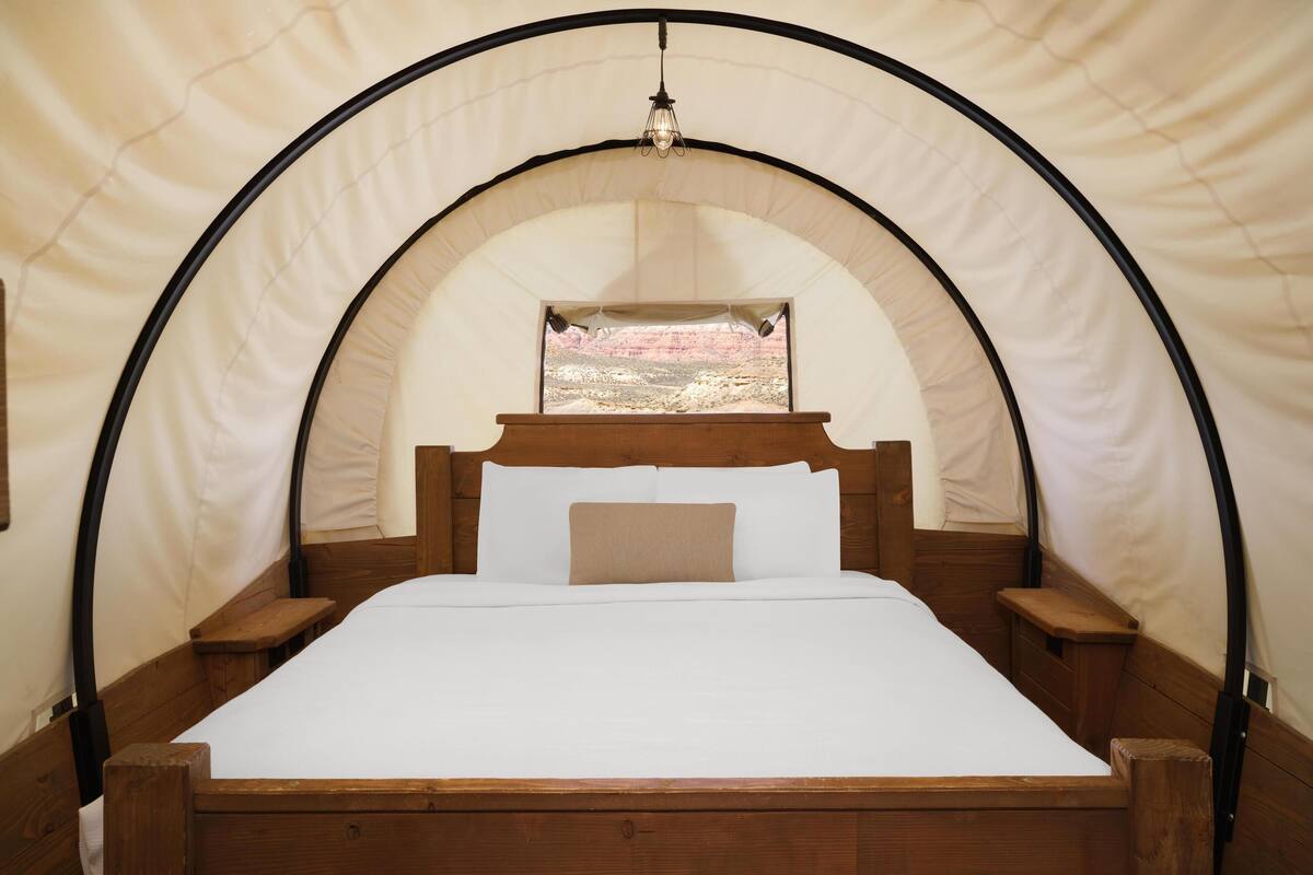 Uma cama com estrutura de madeira e roupa de cama branca, uma mesinha de cabeceira de madeira e uma janela com vista para uma paisagem rochosa.
