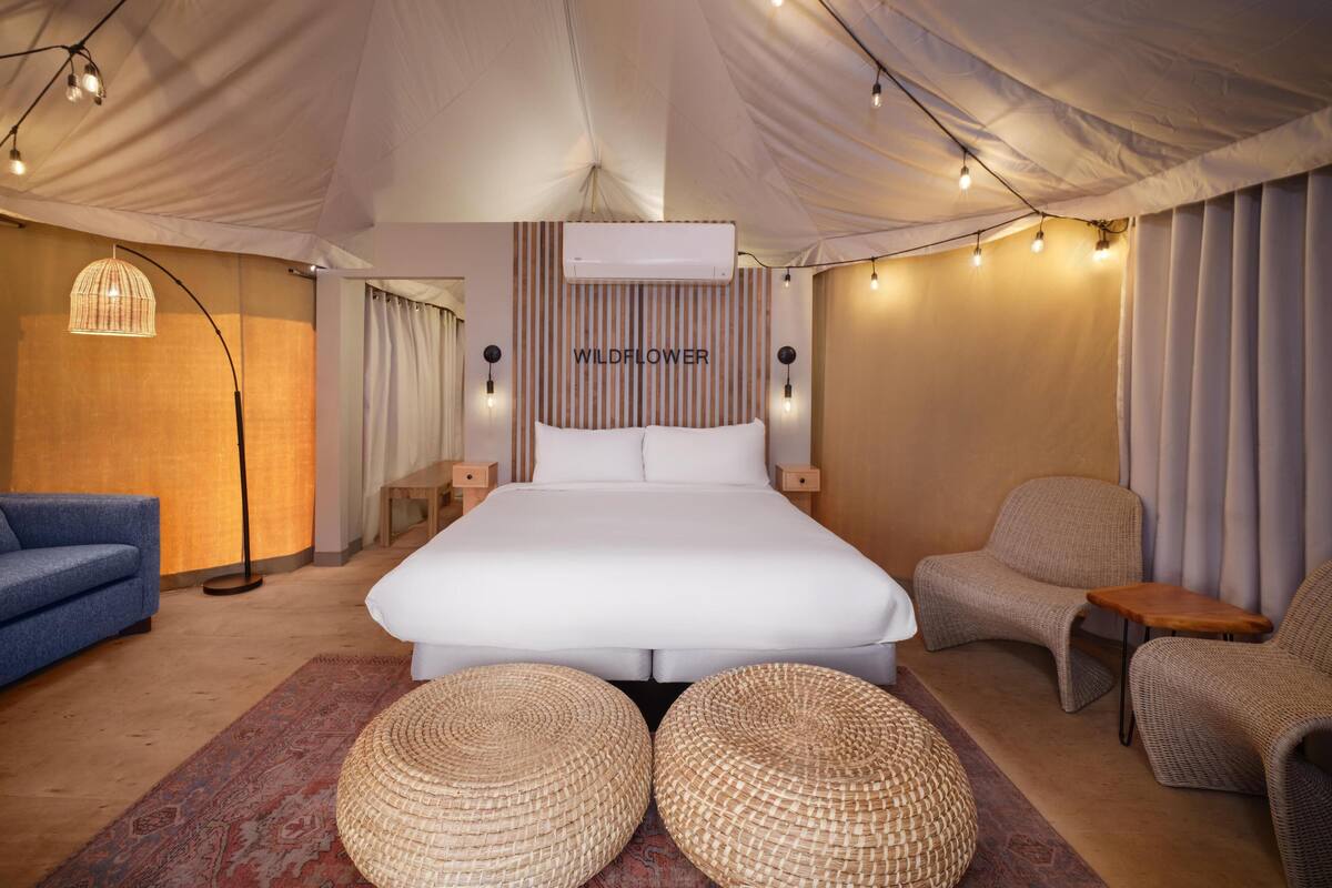 Quarto com uma cama grande, duas mesas de cabeceira, um sofá azul, dois pufes de vime e um ar-condicionado fixado na parede.