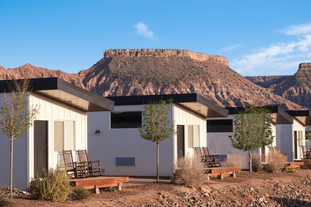 Fileira de cabanas modernas com bancos e portas de madeira, com montanhas ao fundo.