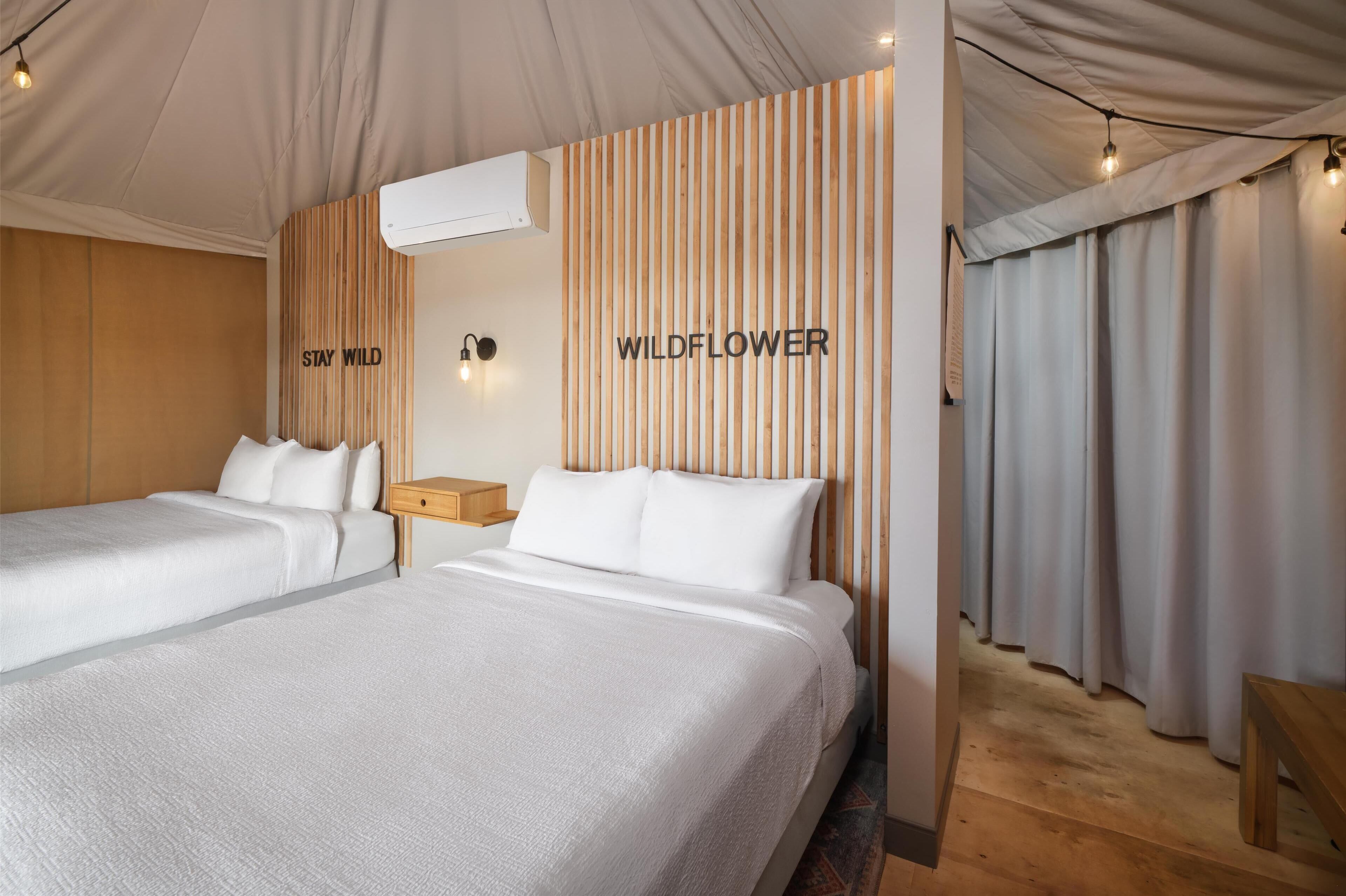 Une chambre d’hôtel avec deux lits, une tête de lit en bois, un climatiseur fixé au mur et une enseigne sur laquelle on peut lire « WILDFLOWER ».