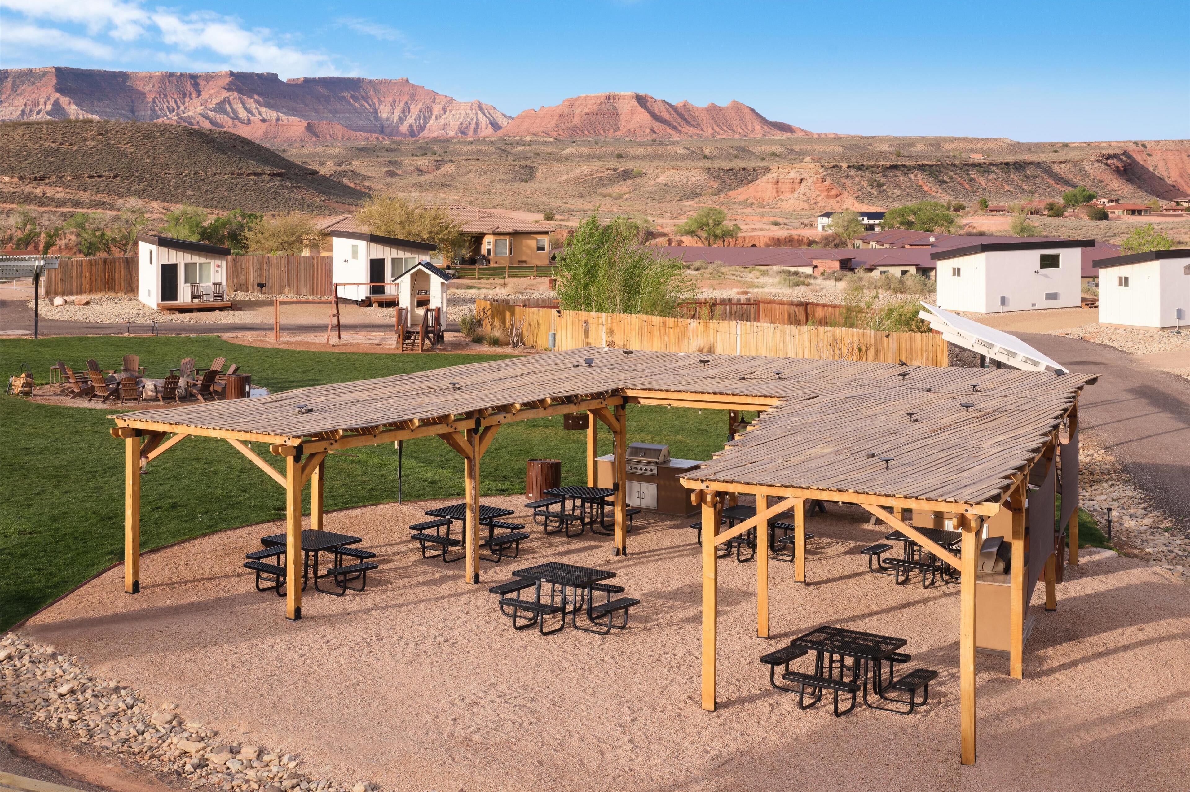 A campground with picnic tables and a covered seating area, surrounded by a wooden fence and houses in a desert landscape.