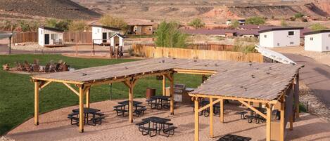 A campground with picnic tables and a covered seating area, surrounded by a wooden fence and houses in a desert landscape.