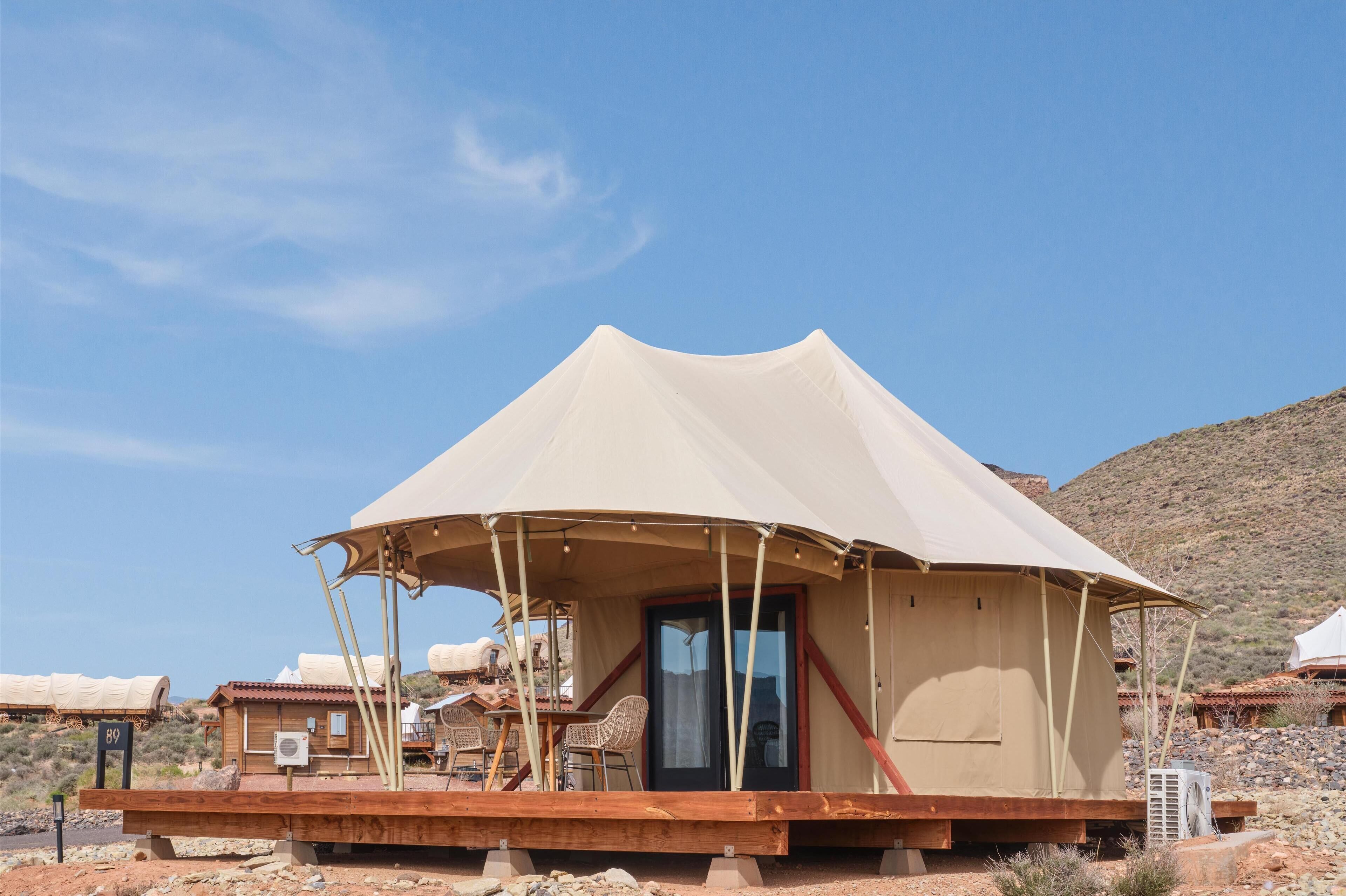 A large, beige tent with a wooden deck and chairs, set in a desert environment with rocky hills in the background.