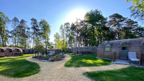 A row of rustic cabins with stone walls and arched windows, surrounded by a forested area and a gravel path. - Iglu Camp Heidewald (Sassenberg)