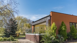 Una casa moderna con un balcón de vidrio, rodeada por un jardín cuidado y altos árboles.