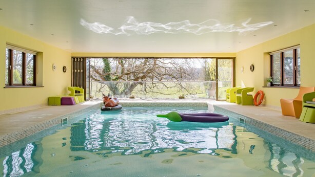 Una piscina interior con paredes de vidrio transparente, vistas a árboles fuera y sillas de colores alrededor de la piscina.