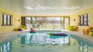 Una piscina interior con paredes de vidrio transparente, vistas a árboles fuera y sillas de colores alrededor de la piscina.