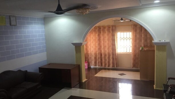 A living room with a sofa, a small table, a ceiling fan, and a view through an archway to a room with curtains. - Nyaboa Hotel (Kumasi)