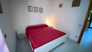 A bedroom with a white bed, a red blanket, a nightstand with a lamp, and three framed pictures on the wall. - Appartamento Rosa La Cinta (San Teodoro)