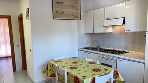 A small kitchen with white cabinets, a gas stove, and a sink. There is a dining table with chairs and a floral tablecloth. - Appartamento Rosa La Cinta (San Teodoro)
