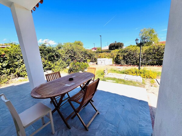 Une terrasse couverte avec une table et des chaises, entourée de verdure et sous un ciel dégagé.