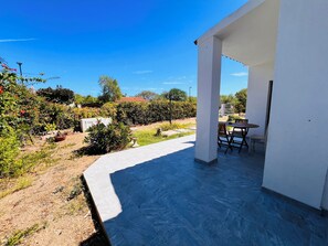 Un espace extérieur abrité, avec une table et des chaises, entouré de verdure et surplombé d’un ciel bleu dégagé.