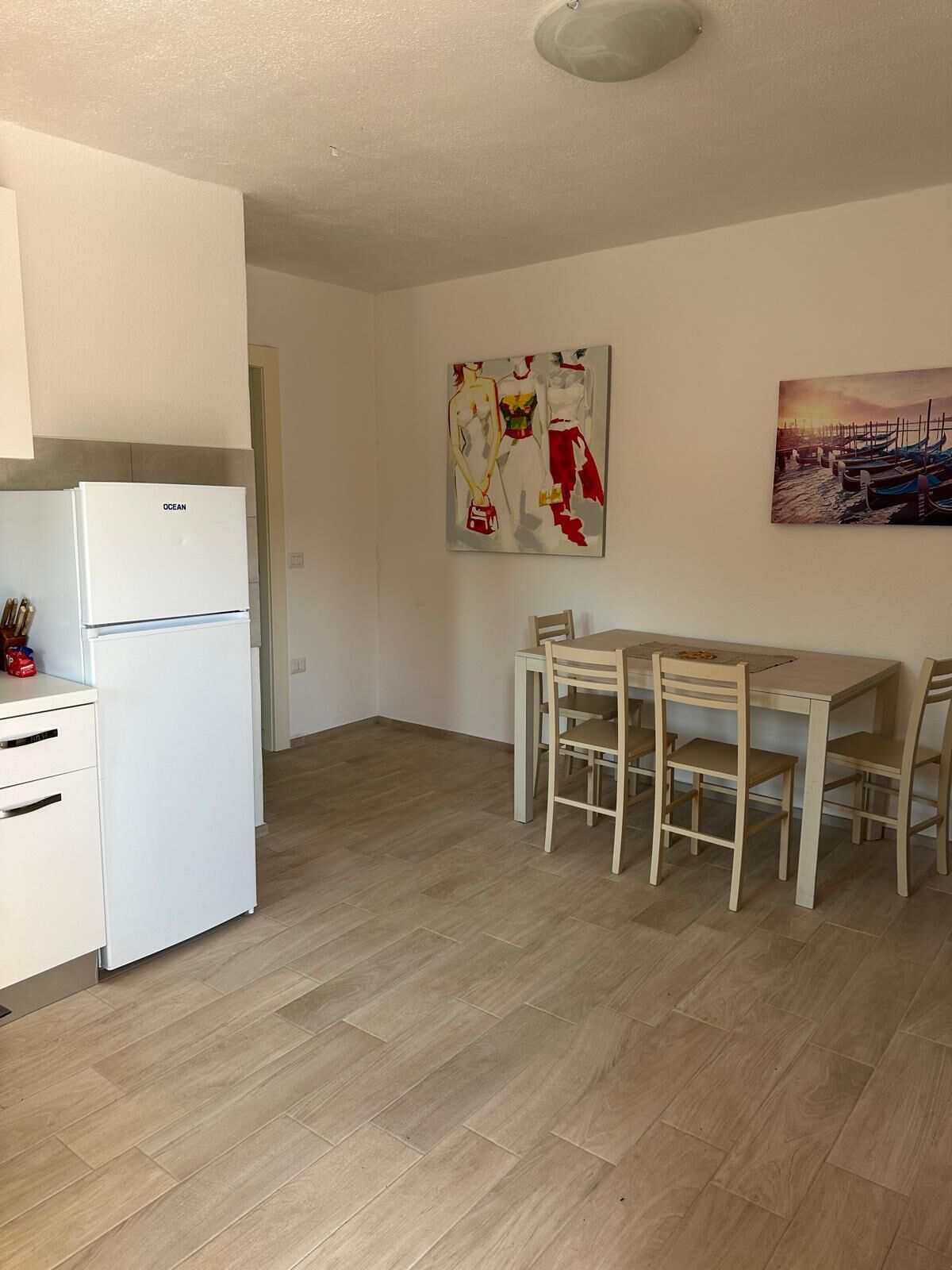 Une cuisine avec un réfrigérateur blanc, une table à manger en bois et quatre chaises, ainsi que deux tableaux accrochés au mur.